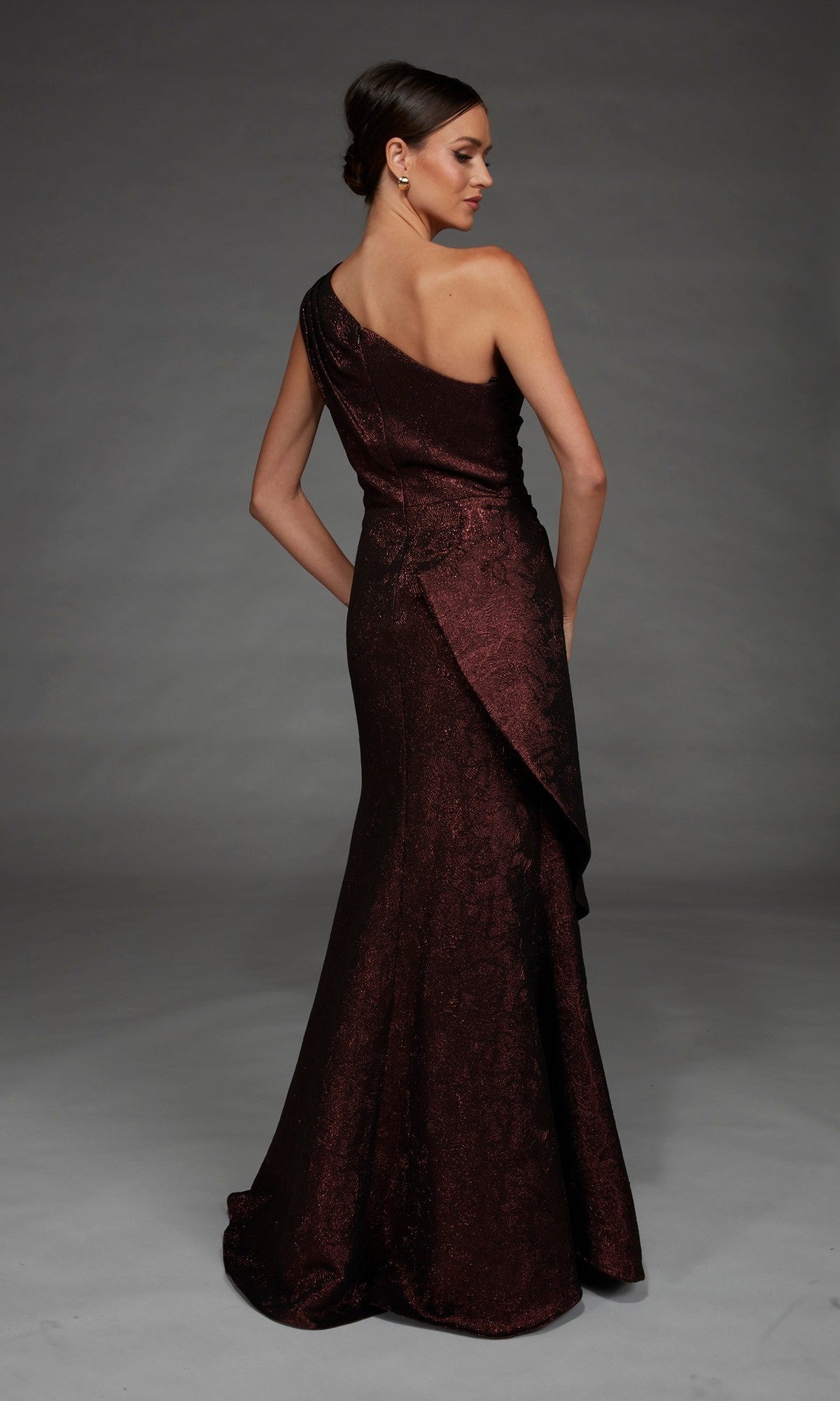 A woman stands facing away against a plain gray background, wearing the Long Formal Dress: Alyce 27885—a dark, shimmery, one-shoulder evening gown crafted from elegant metallic jacquard fabric that catches the light with every subtle movement.
